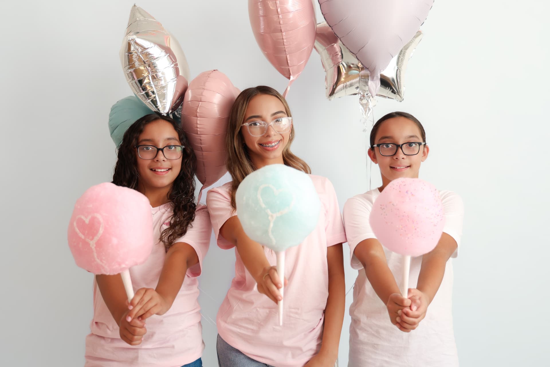 Three kids holding heart-stamped cotton candy puffs with pastel balloons